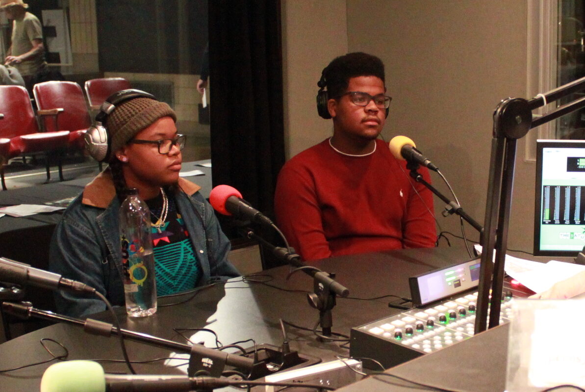 Two teens wearing headphones sitting behind microphones in a radio studio