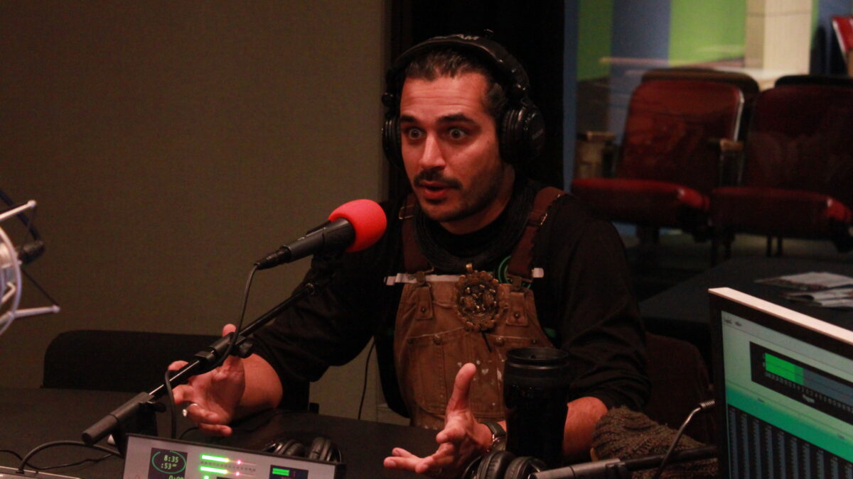 Adult wearing brown overalls and headphones speaking into a microphone and gesturing with their hands in a radio studio