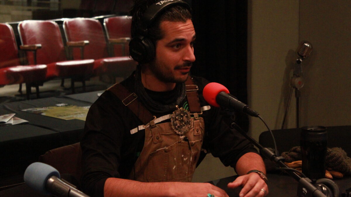Adult wearing brown overalls and headphones speaking into a microphone in a radio studio
