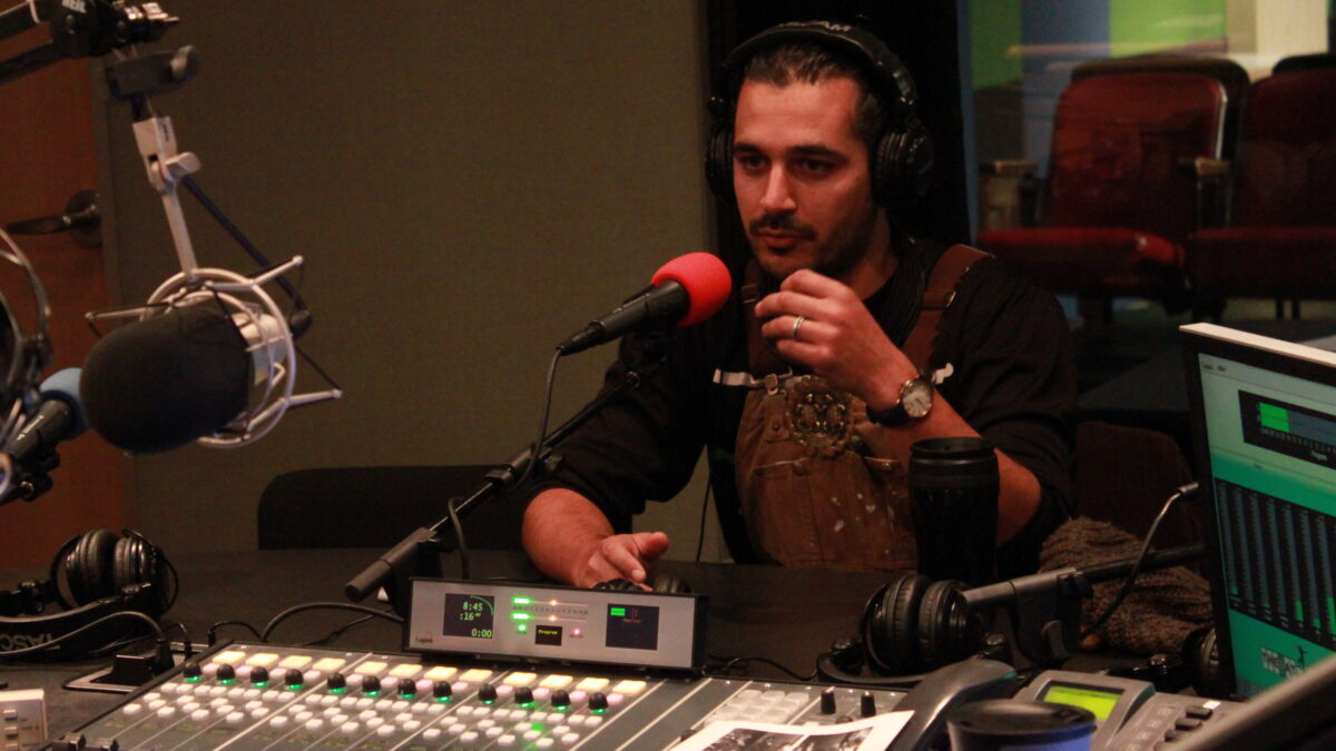 Adult wearing brown overalls and headphones speaking into a microphone and gesturing with their hands in a radio studio