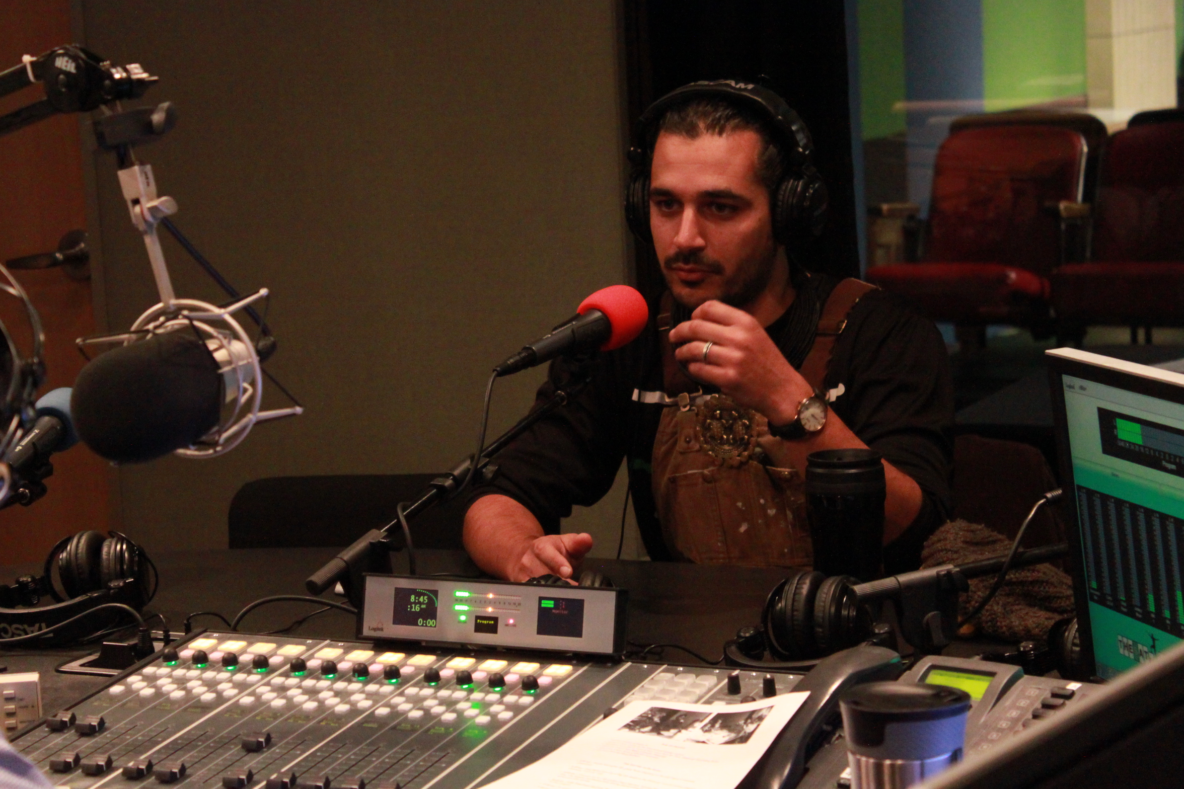 Adult wearing brown overalls and headphones speaking into a microphone and gesturing with their hands in a radio studio