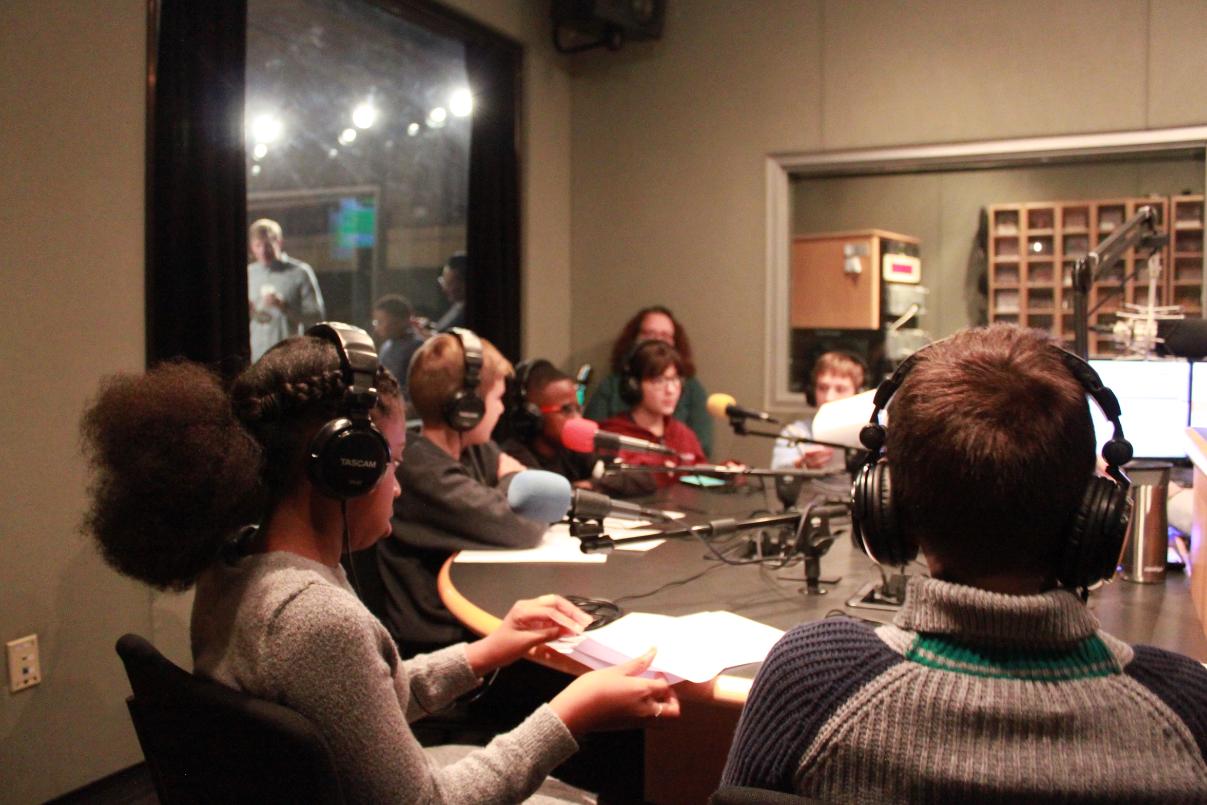 Six youth wearing headphones sitting around a table in a radio studio while an adult supervises