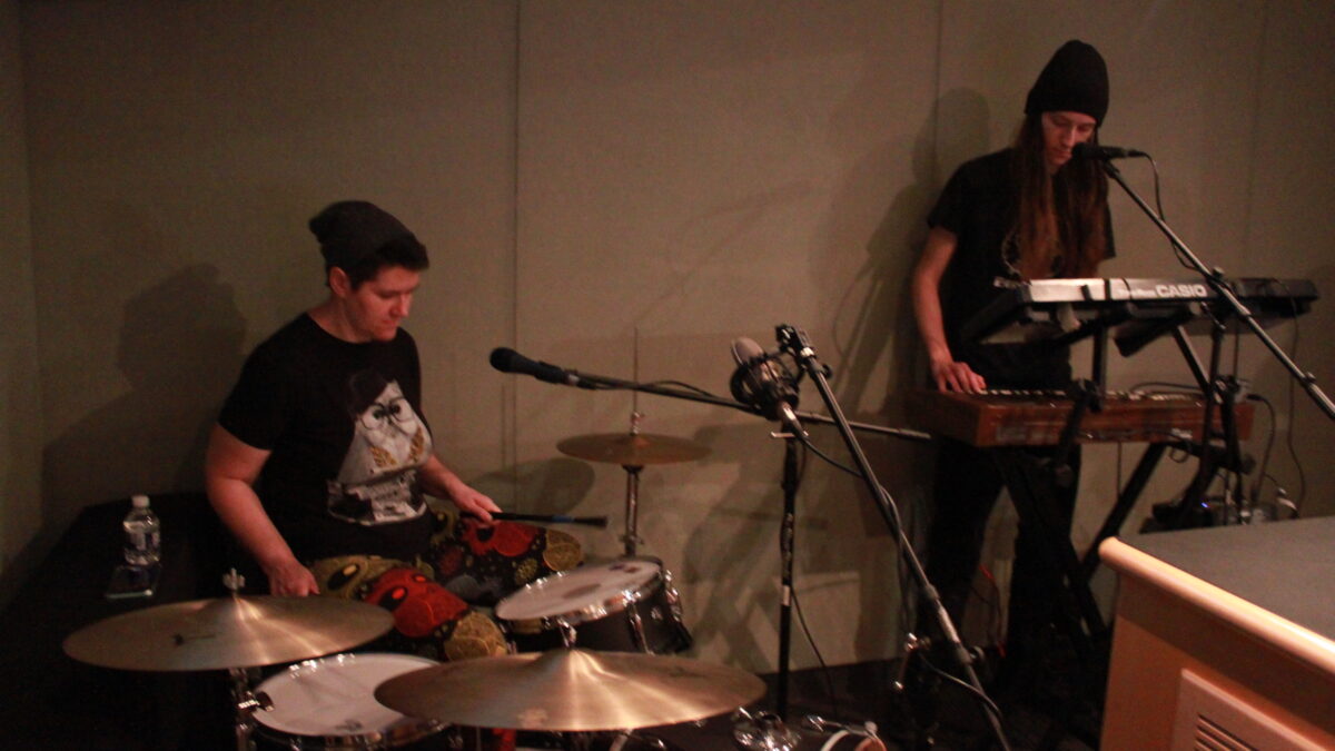 An adult playing on a drum set in front of microphones and an adult playing synthesizers in front of a microphone in a radio studio