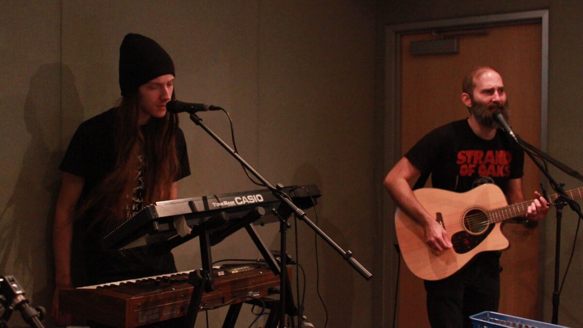 An adult playing synthesizers and an adult playing guitar and singing into a microphone in a radio studio