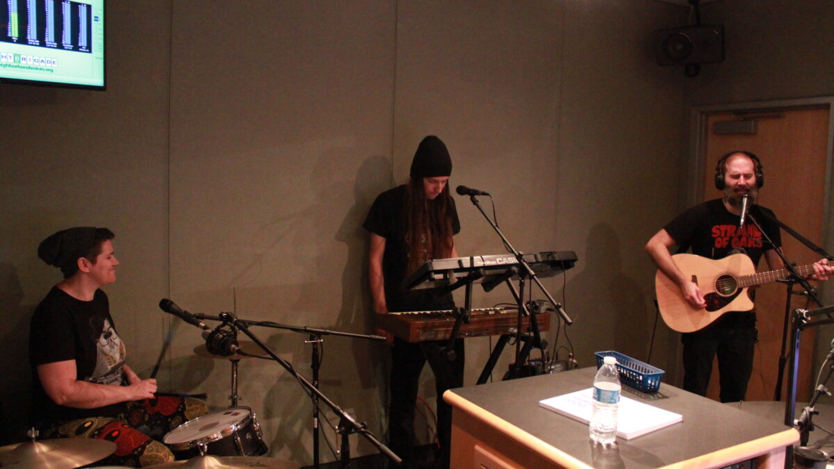 An adult playing on a drum set, an adult playing on two synthesizers, and an adult playing guitar and singing into a microphone in a radio studio