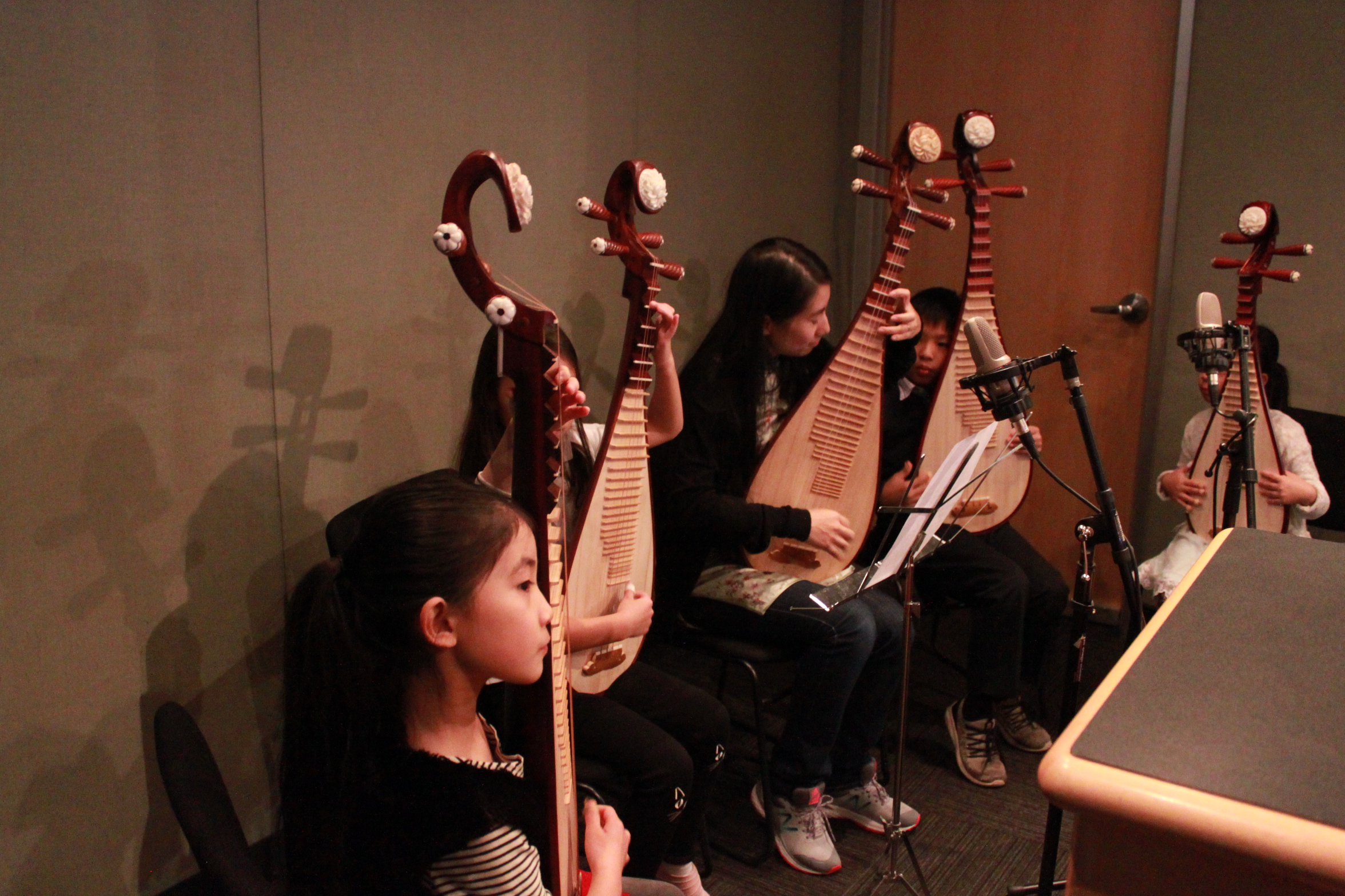 Four children and one adult holding ethnic stringed instruments and looking at sheet music on a music stand in a radio studio