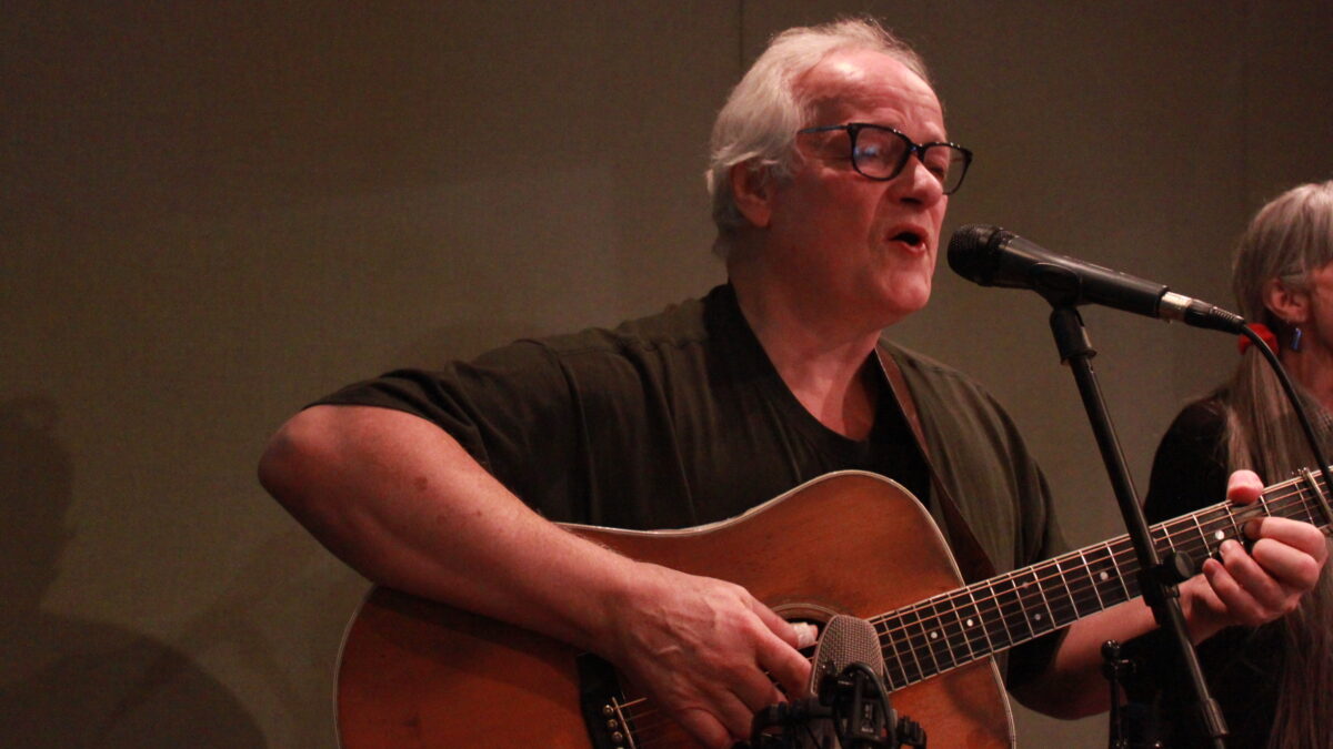 Adult playing guitar and singing into a microphone in a radio studio