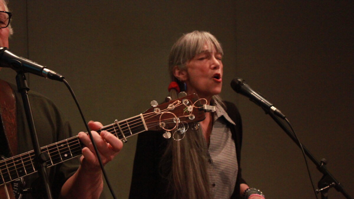 Two adults singing into microphones in a radio studio, one playing guitar