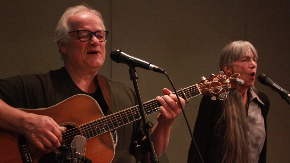 Two adults singing into microphones in a radio studio, one playing guitar