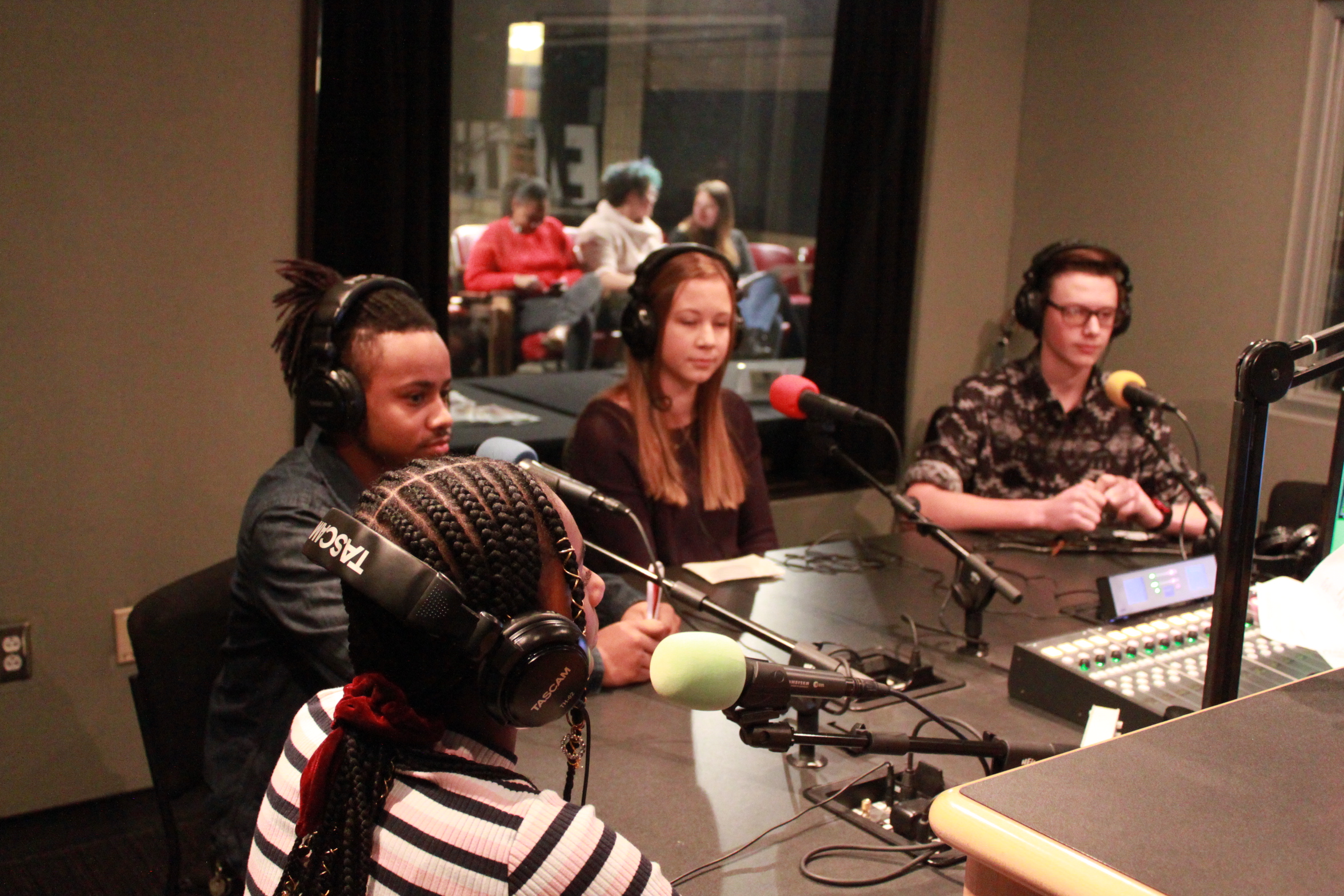 Four teens wearing headphones sitting behind microphones in a radio studio