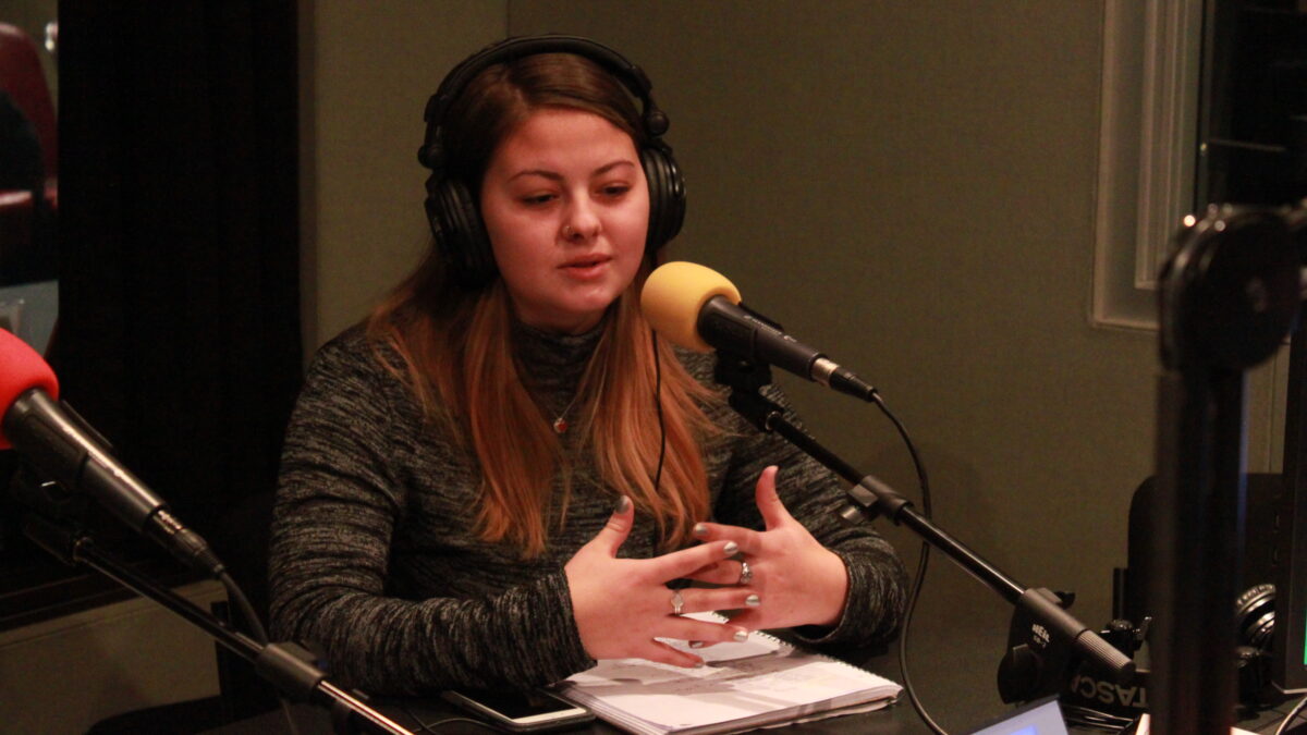 Teen wearing headphones speaking into a microphone and gesturing with their hands in a radio studio