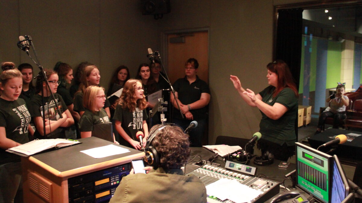 Eleven youth singing in a choir while an adult conducts and two other adults watch in a radio studio