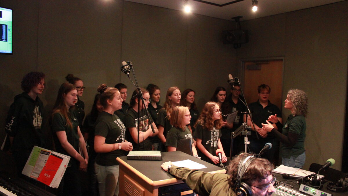 Fifteen youth singing in a choir while an adult conducts them and another adult listens in a radio studio