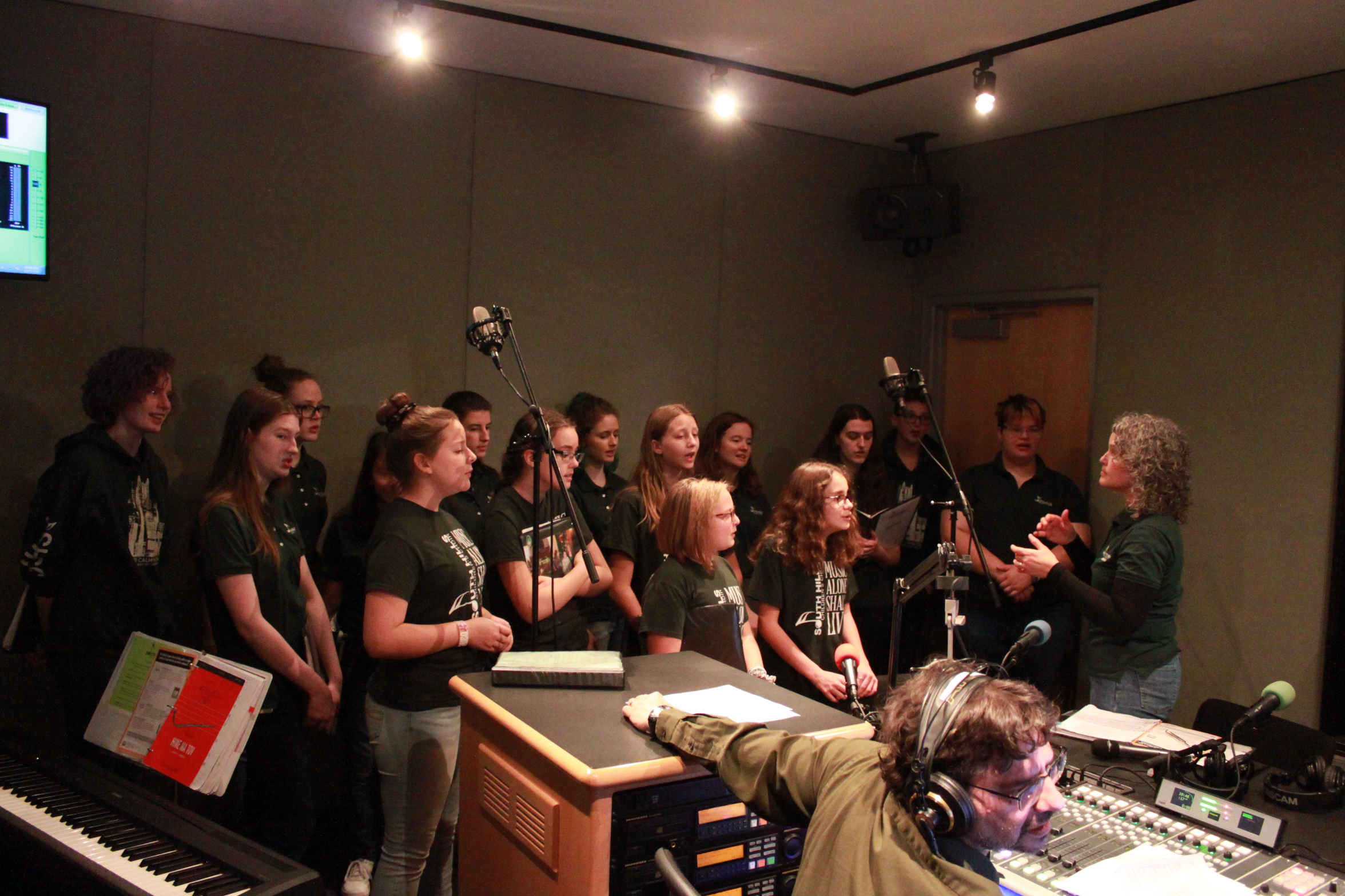 Fifteen youth singing in a choir while an adult conducts them and another adult listens in a radio studio
