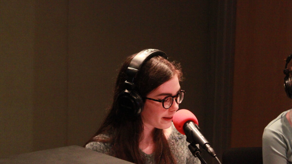 Individual wearing glasses and headphones speaking into a microphone in a radio studio