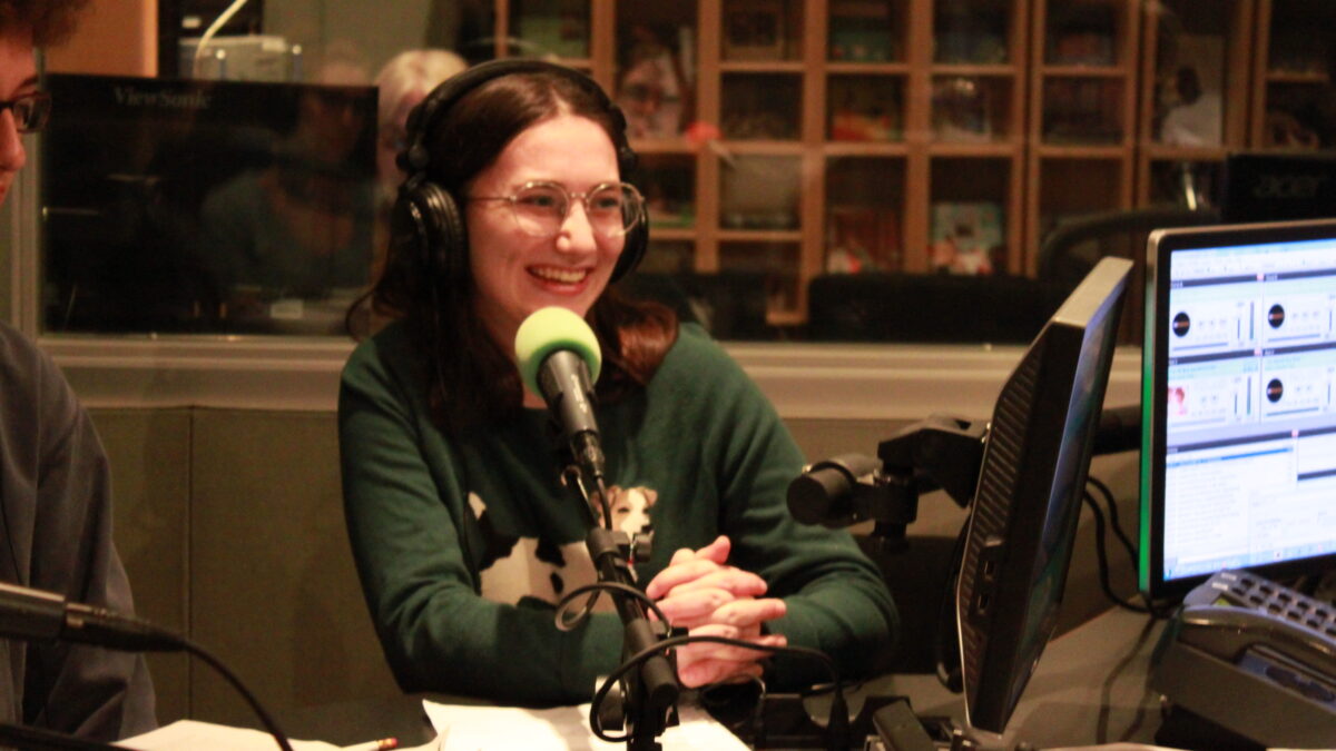 Individual wearing glasses and headphones speaking into a microphone in a radio studio