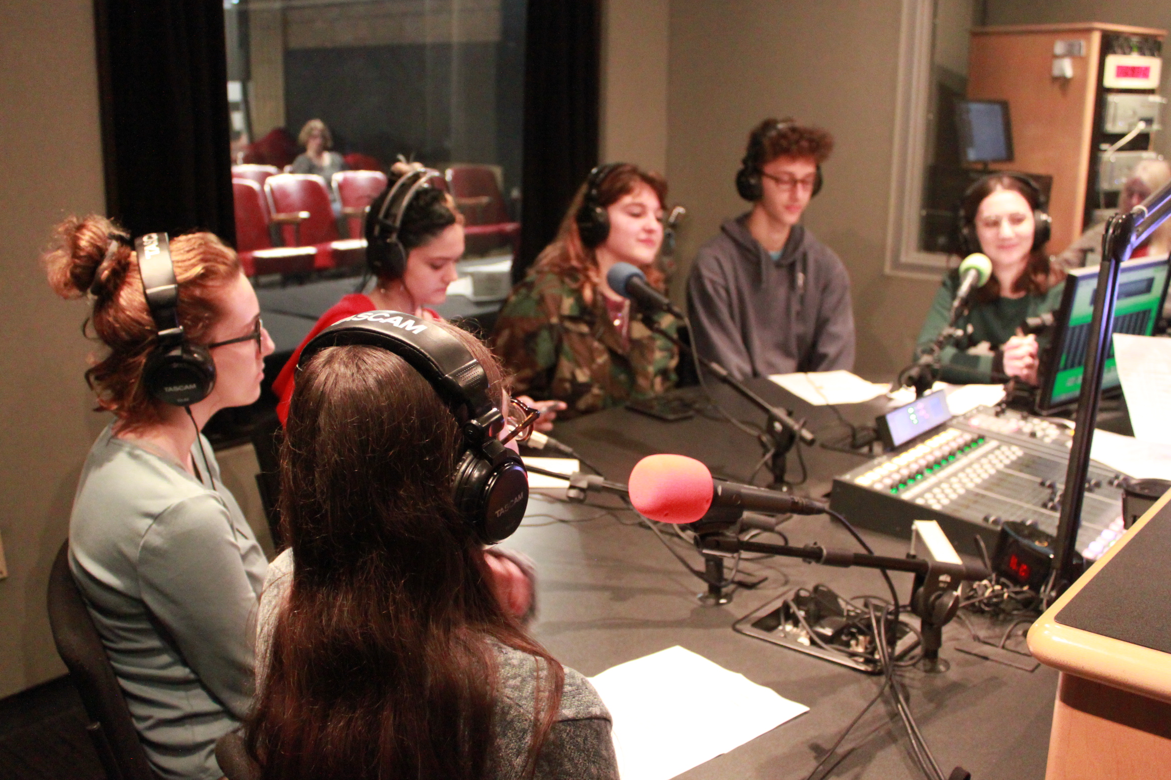 Six teens wearing headphones sitting in front of microphones in a radio studio
