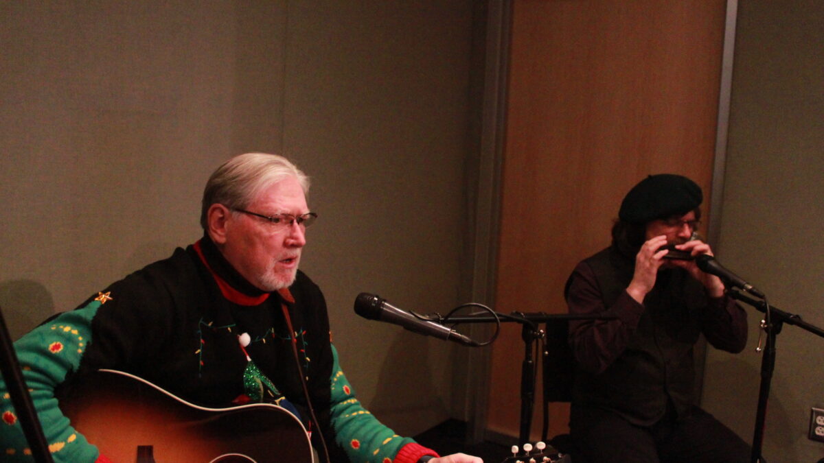 Adult playing guitar and singing into a microphone and an adult playing harmonica into a microphone in a radio studio