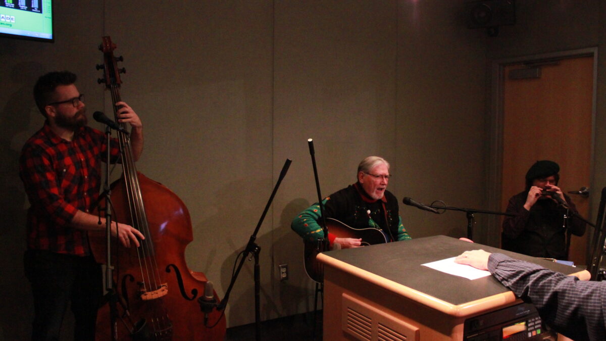 An adult playing upright bass, an adult playing guitar and singing into a microphone, and an adult playing harmonica into a microphone in a radio studio