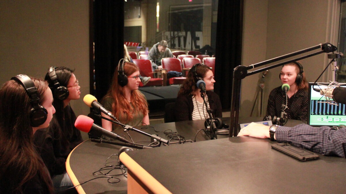 Five teens wearing headphones and speaking into microphones in a radio studio