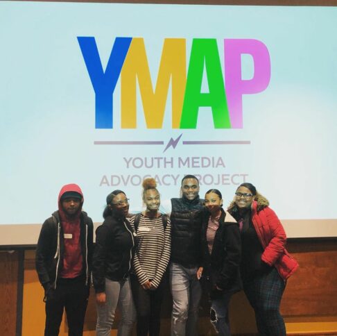 Six teens standing beneath a projector displaying a large blue letter Y, a large orange letter M, a large light green letter A, a large purple letter P, and the text "Youth Media Advocacy Project" in black