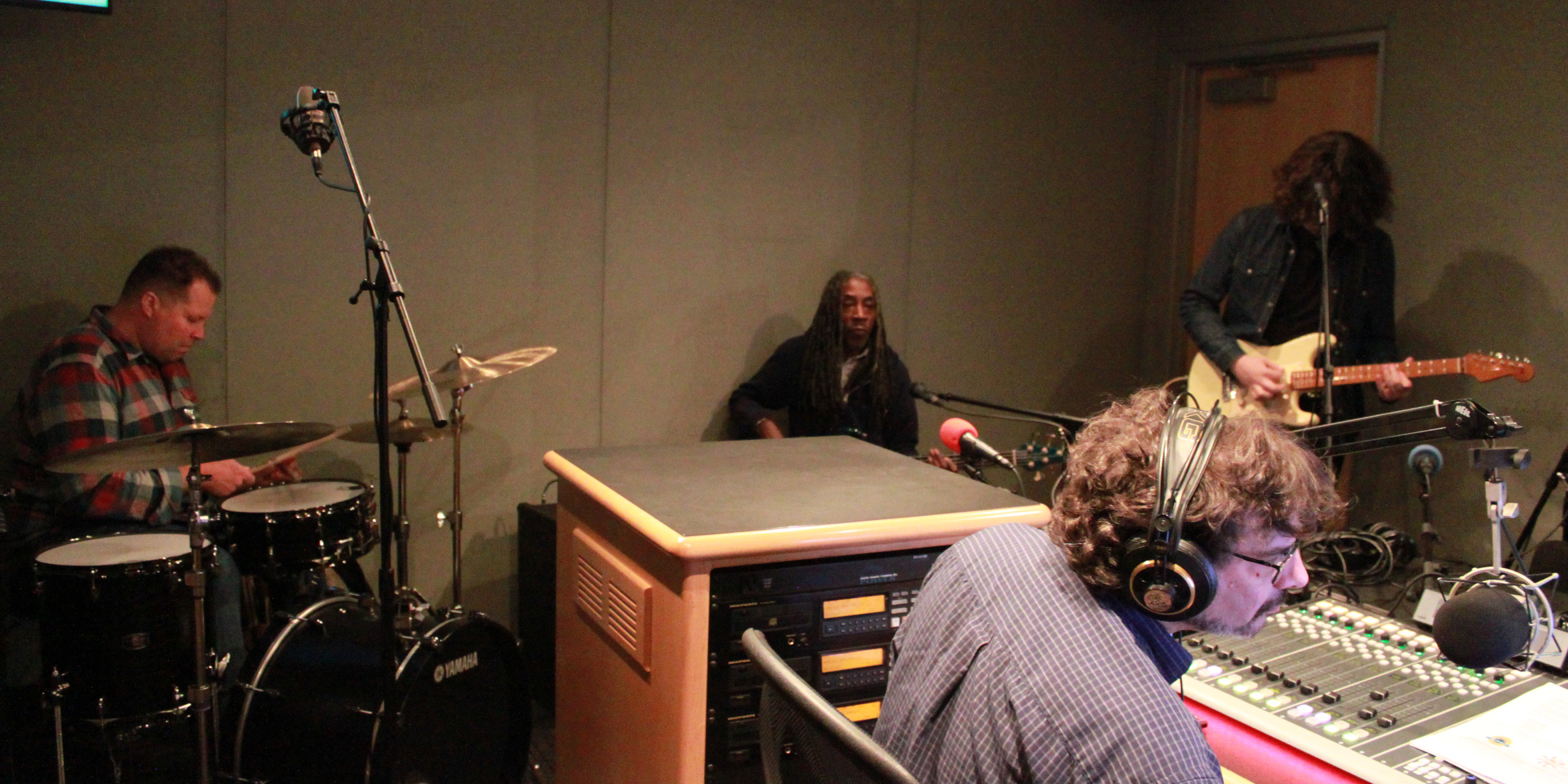 Three adult musicians performing in a radio studio, one on drums, one on bass, one on guitar
