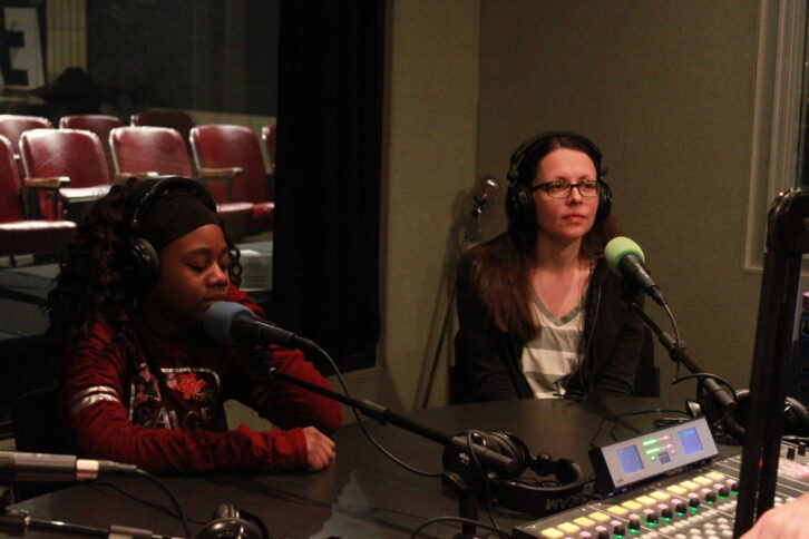 Two people wearing headphones sitting in front of microphones in a radio studio