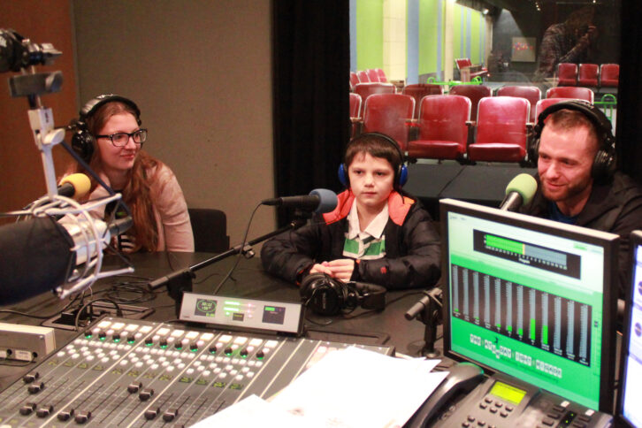 Two people and one child wearing headphones sitting in front of microphones in a radio studio