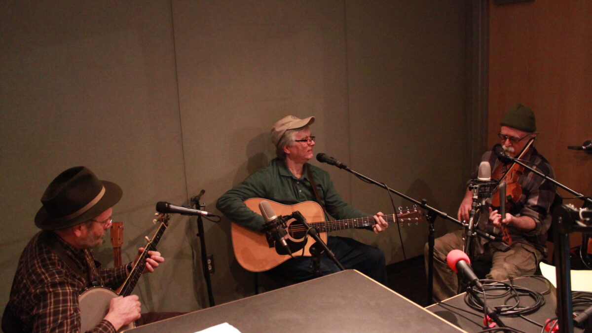 Three adults sitting in a radio studio in front of microphones, one playing banjo, one playing guitar, one playing violin