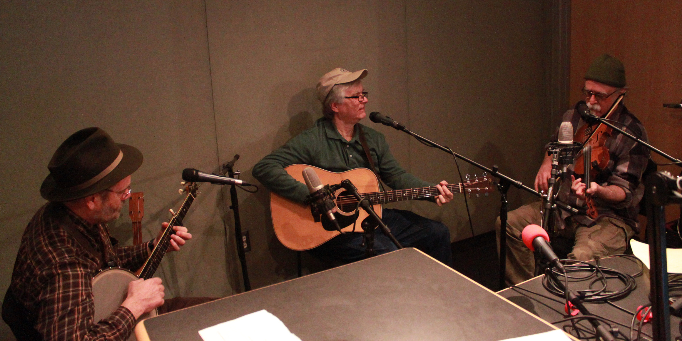 Three adults sitting in a radio studio in front of microphones, one playing banjo, one playing guitar, one playing violin
