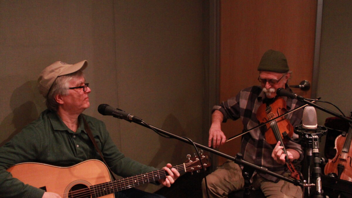 An adult playing guitar and an adult playing violin in front of microphones in a radio studio