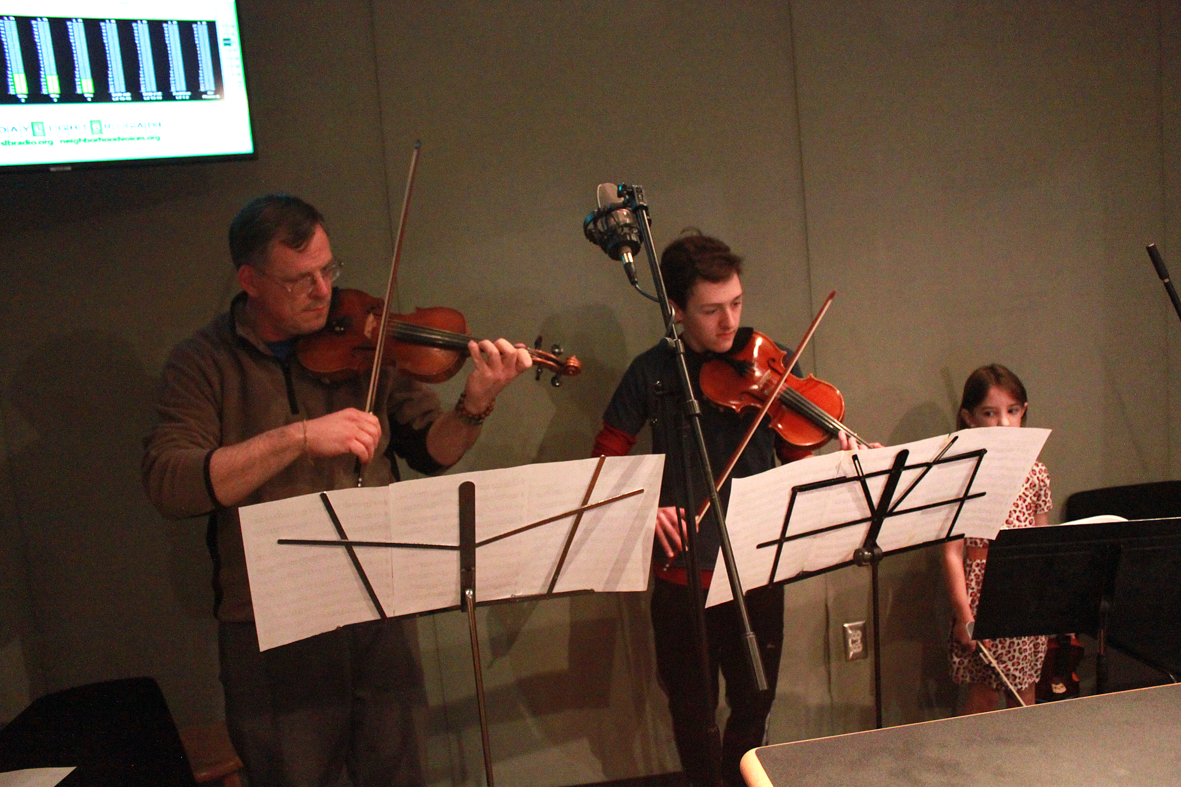 An adult and a youth playing violins and looking at sheet music on music stands while a child holding a violin watches in a radio studio