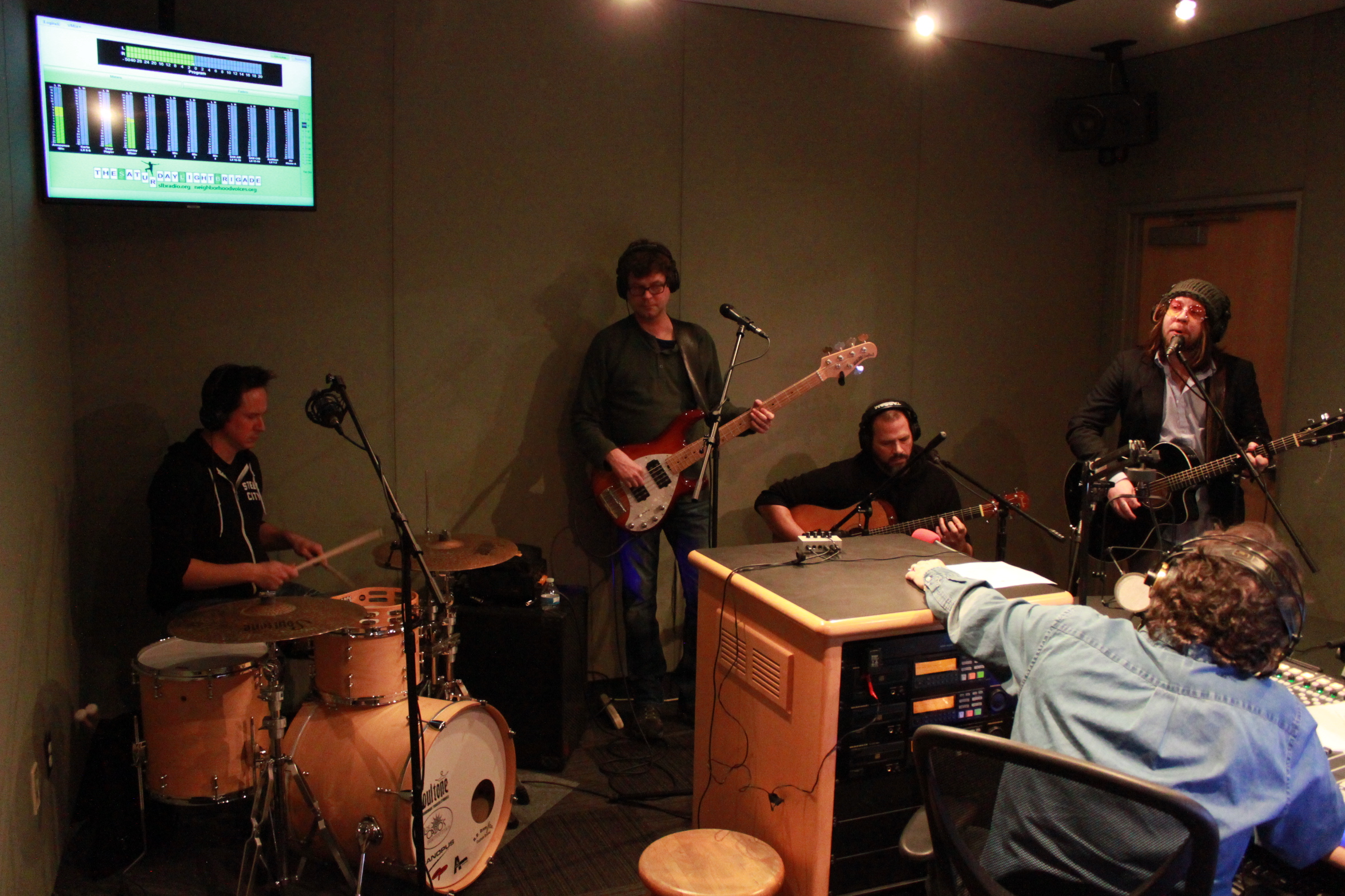 Four adults wearing headphones, one playing a drum set, one playing bass guitar, two playing guitar, and one singing into a microphone, with another adult wearing headphones listening in a radio studio
