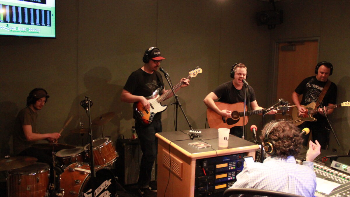 Four adults wearing headphones performing on drum set, bass guitar, acoustic guitar, and electric guitar in a radio studio, while another adult wearing headphones is listening