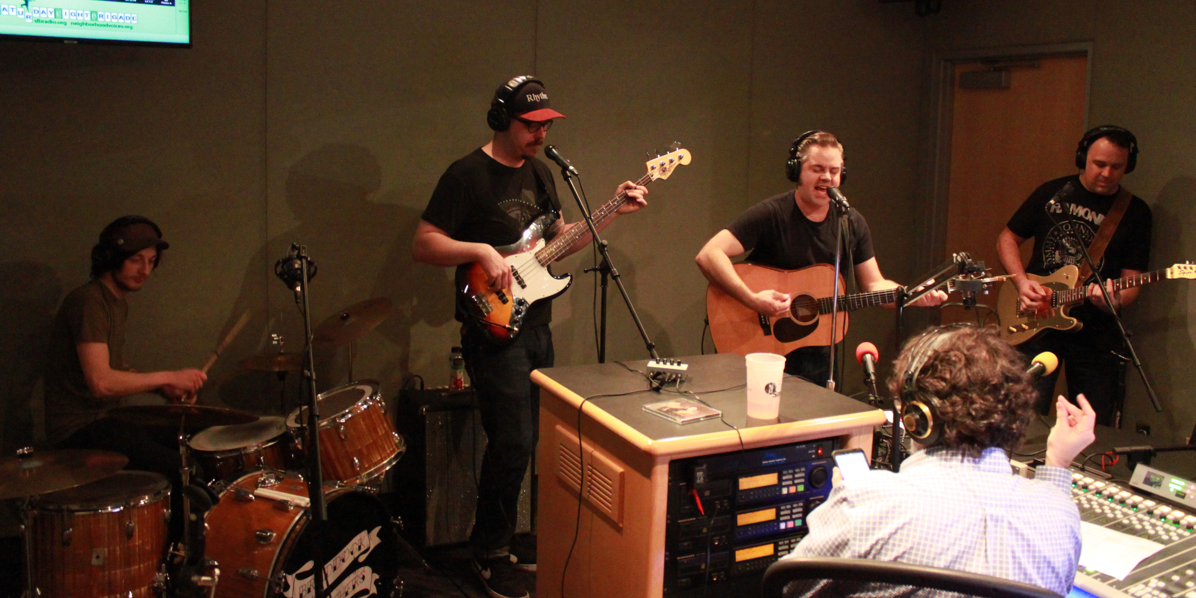 Four adults wearing headphones performing on drum set, bass guitar, acoustic guitar, and electric guitar in a radio studio, while another adult wearing headphones is listening