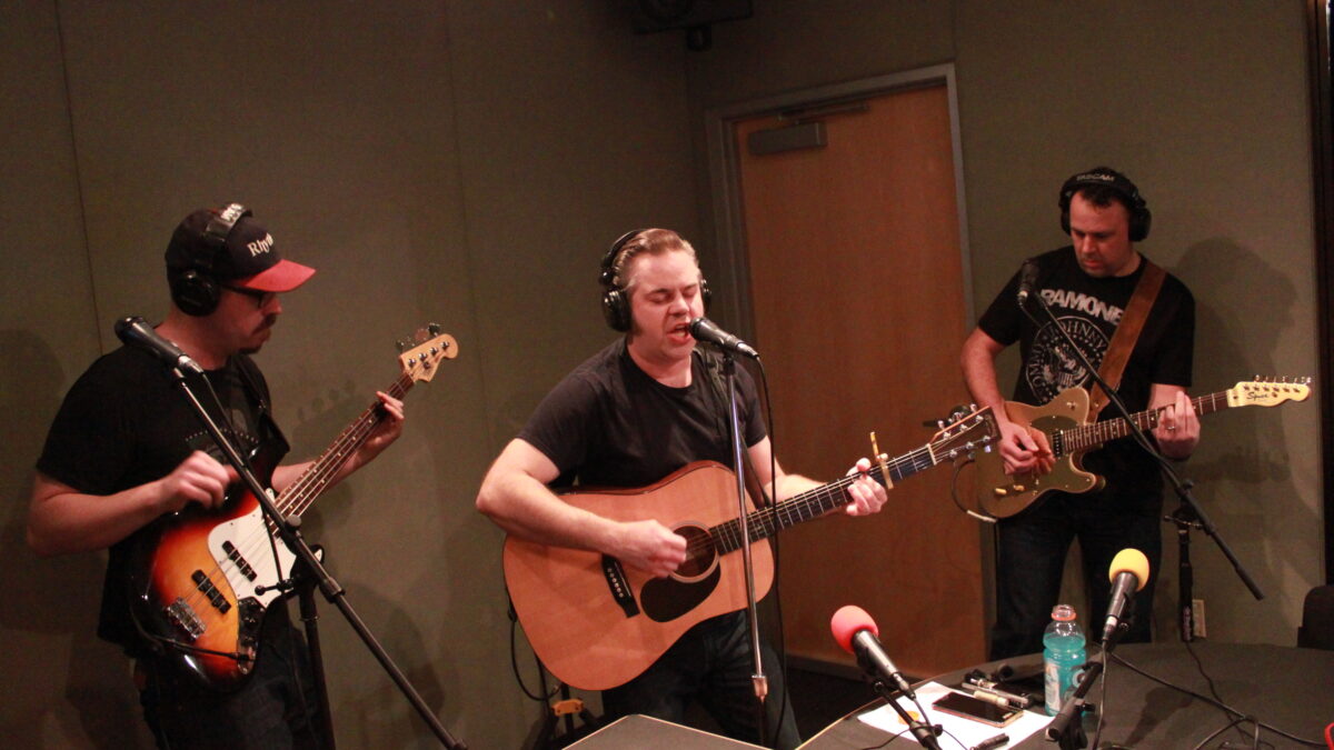 Three adults wearing headphones, one playing bass guitar, one playing electric guitar, and one playing acoustic guitar and singing into a microphone, all in a radio studio
