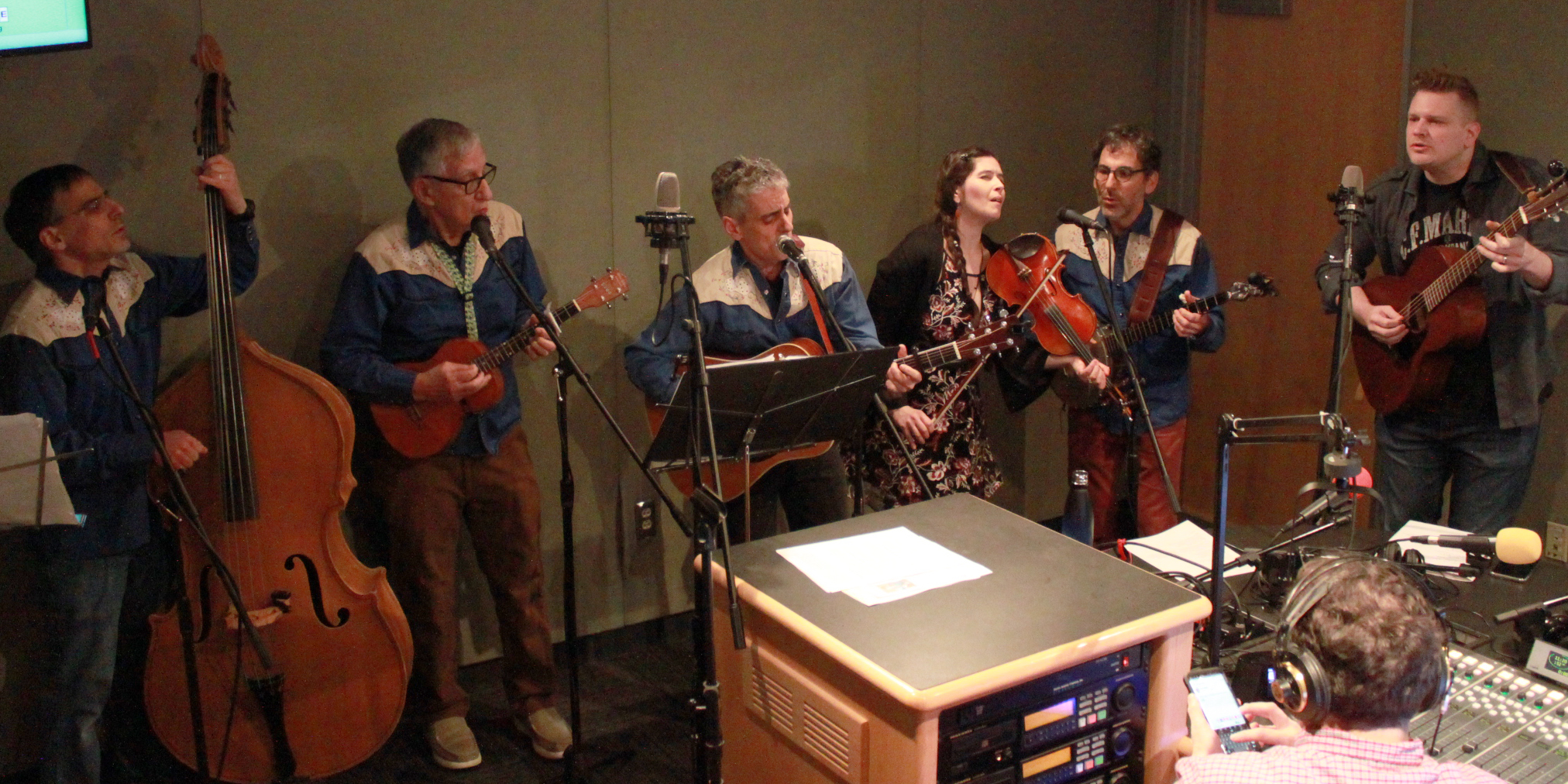 Six adults singing into microphones in a radio studio as another adult wearing headphones listens, one adult playing upright bass, one adult playing ukulele, two adults playing guitar, one adult playing violin, and another playing banjo