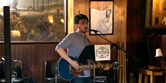 Adult playing a guitar and singing into a microphone in front of a wooden wall with a painting of an old war