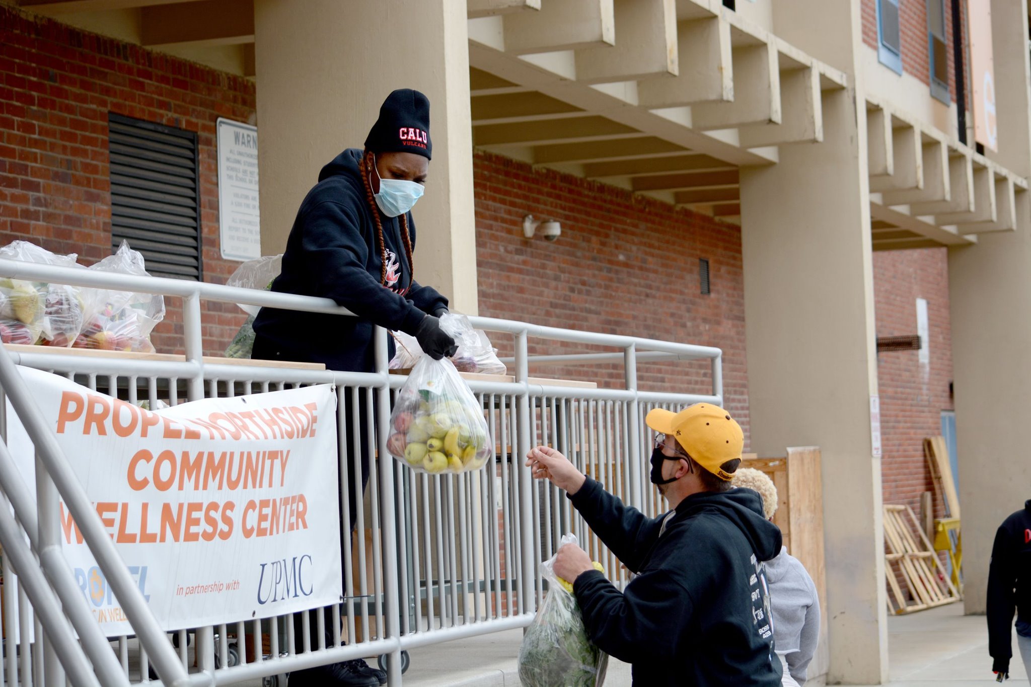 An adult in a face mask handing a bag of fruit to another adult in a face mask over a gray handrailing in front of a brick wall