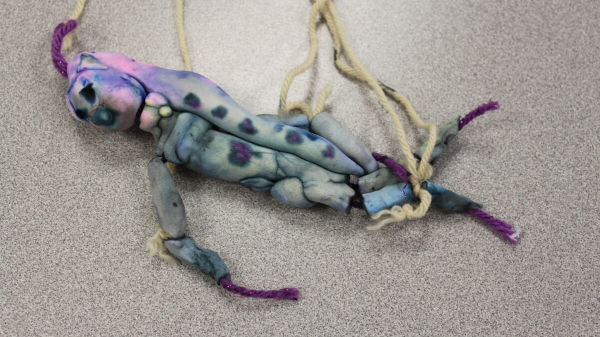 A sculpture of an individual made with light blue clay and white and purple strings