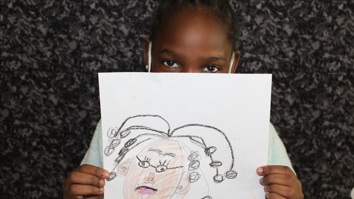 A child holding up a piece of paper with a drawing of a brown face with glasses and long hair