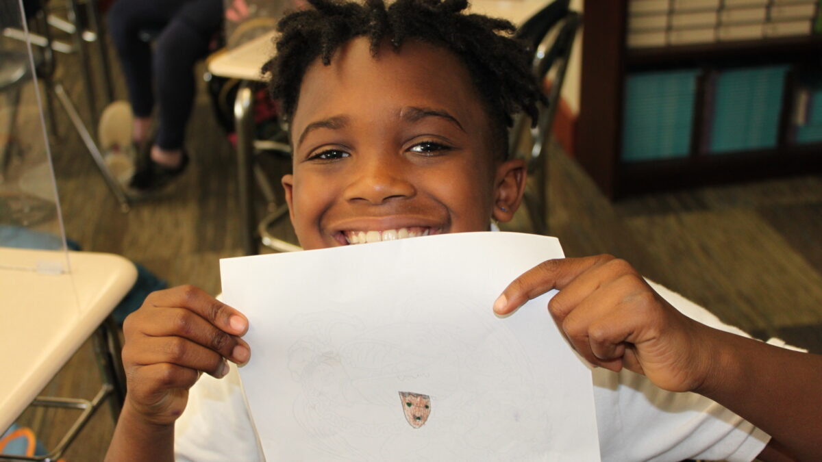 A child smiling and holding up a piece of paper with a small brown face drawn onto it