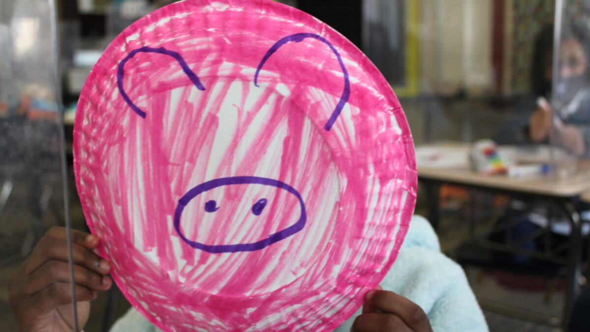 A child holding up a pink paper plate, with pig ears and a pig nose drawn on with a blue marker