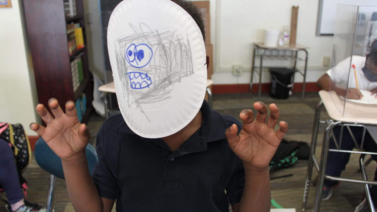 A child with their face covered by a paper plate with large blue eyes, a blue mouth filled with sharp teeth, and gray scribbles drawn on