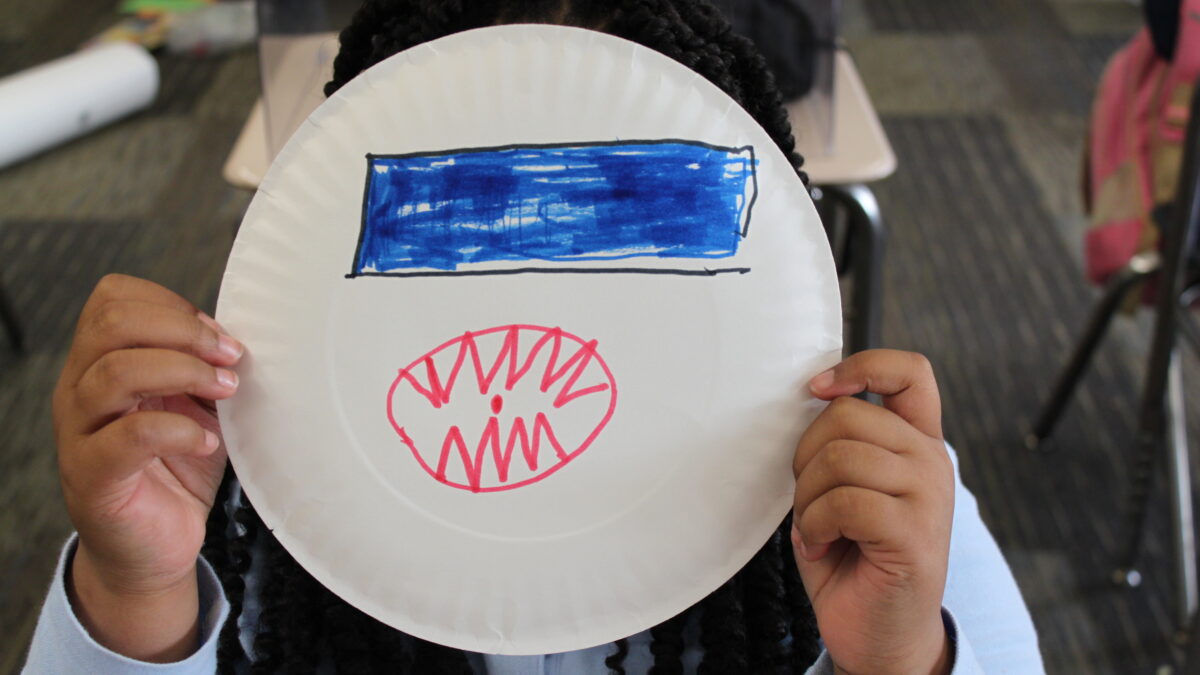 A child holding up a paper plate with a blue rectangle and a red mouth filled with sharp teeth drawn on