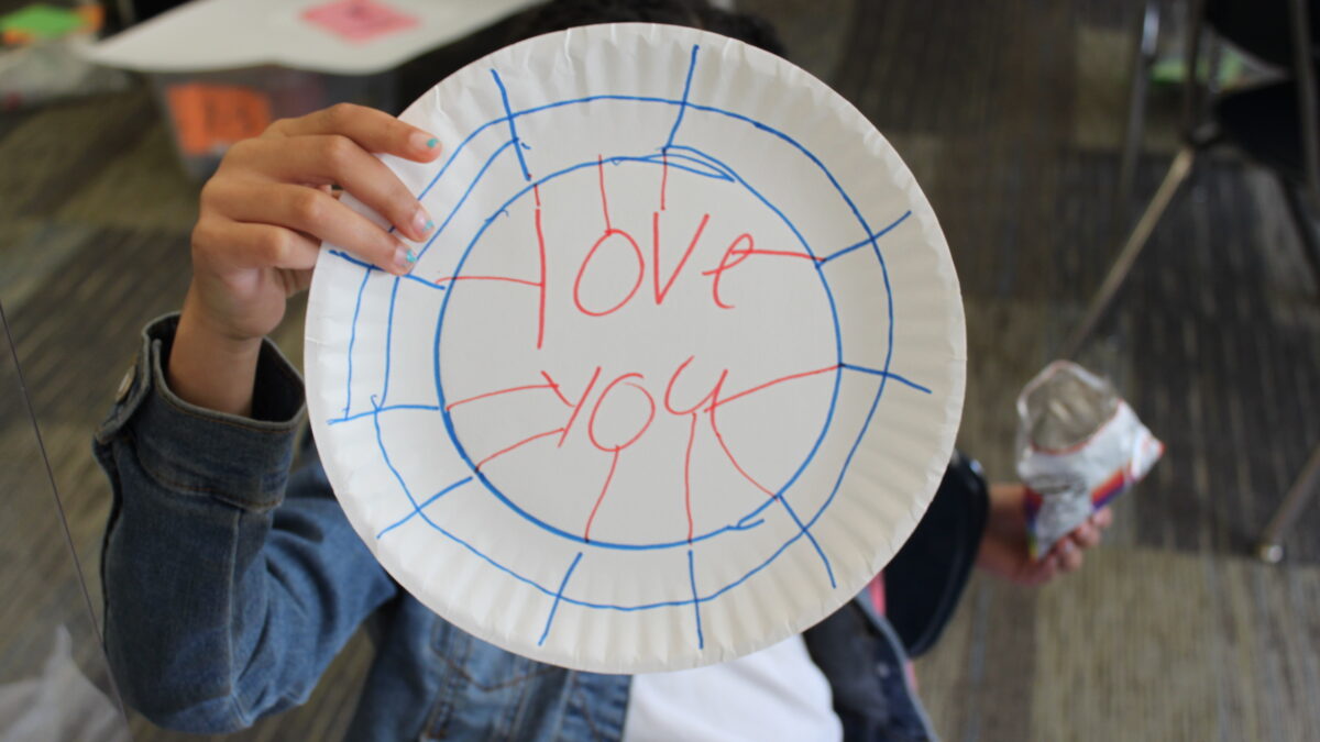 A child holding up a paper plate with a blue drawn on border, and the text "love you" written in red in the center