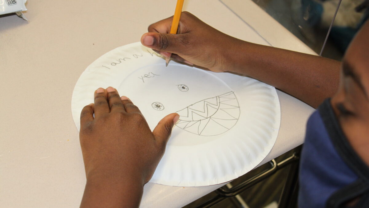 A child drawing onto a paper plate that already has the text "I am a hacker yea" written on it, and two bloodshot eyes and a mouth full of sharp teeth drawn onto the plate