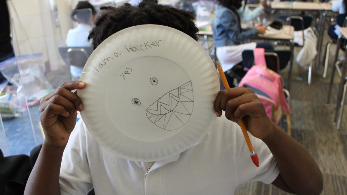 A child holding a paper plate with the text "I am a hacker yes" written on it, as well as two bloodshot eyes and a mouth full of sharp teeth drawn onto the plate