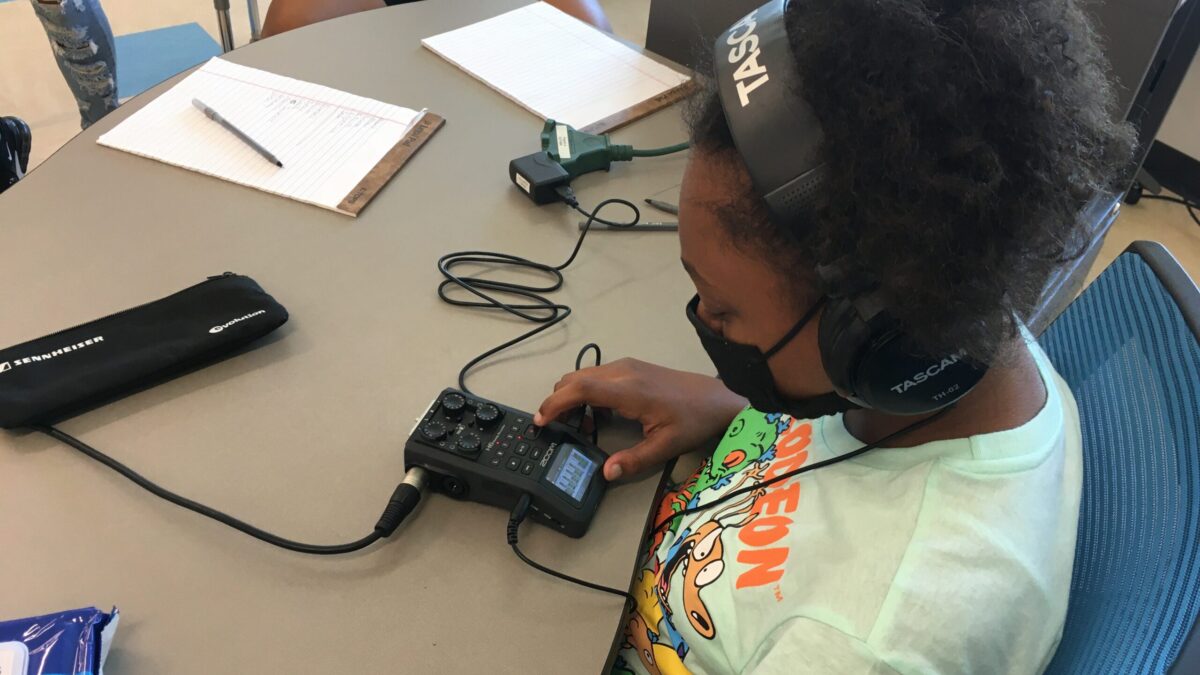 Youth wearing headphones and a face mask interacting with a portable recorder on a desk