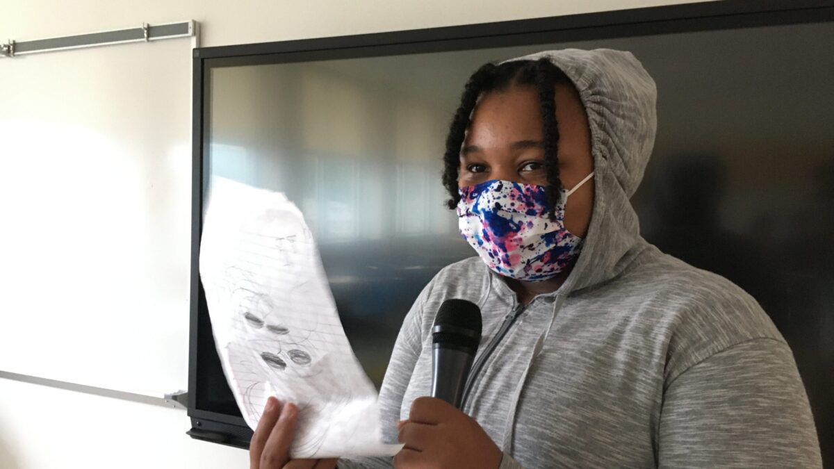 Youth wearing a face mask speaking into a microphone and looking at a piece of notebook paper